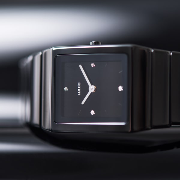 RADO © 2025 Eine elegante, schwarze, rechteckige Armbanduhr mit einem Metallarmband. Die Uhr verfügt über ein minimalistisches Design mit drei diamantenähnlichen Punkten und schlanken, silbernen Zeigern auf dem Zifferblatt und bietet ein modernes, elegantes Erscheinungsbild.