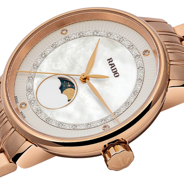 RADO © 2025 Eine luxuriöse Armbanduhr aus der Rado Coupole Kollektion mit Gehäuse und Armband aus Roségold. Das Zifferblatt ist mit Perlmutt-Finish, Diamant-Stundenmarkierungen, goldfarbenen Zeigern und einer Mondphasenanzeige auf der 6-Uhr-Position versehen. Der Markenname „RADO“ ziert das Zifferblatt.
