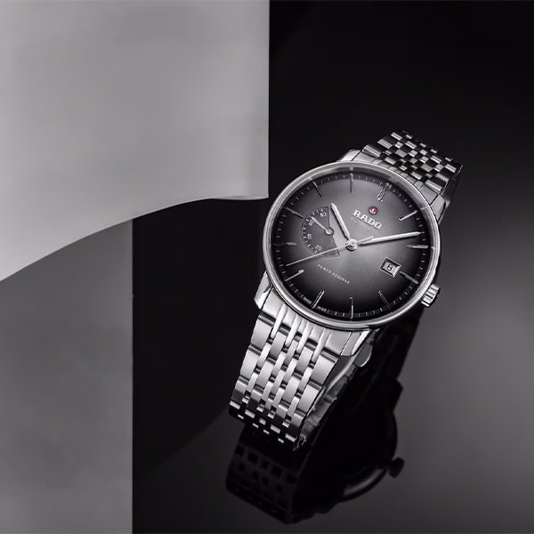 RADO © 2025 Eine elegante, silberne Rado-Armbanduhr aus der Rado Coupole Kollektion mit schwarzem Zifferblatt wird auf einer reflektierenden schwarzen Oberfläche präsentiert. Die Uhr verfügt über ein Datumsfenster, ein Sekunden-Hilfszifferblatt und ein Edelstahlarmband vor einem Hintergrund aus glatten, geschwungenen weißen und schwarzen Elementen.