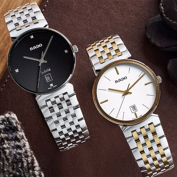 RADO © 2025 Zwei Uhren aus der Rado Florence Kollektion liegen auf einer Holzoberfläche. Die linke hat ein schwarzes Zifferblatt mit silbernen Zeigern und Markierungen, eine Datumsanzeige und ein silbernes Armband. Die rechte hat ein weißes Zifferblatt mit goldenen Zeigern und Markierungen, eine Datumsanzeige und ein zweifarbiges Armband aus Silber und Gold.