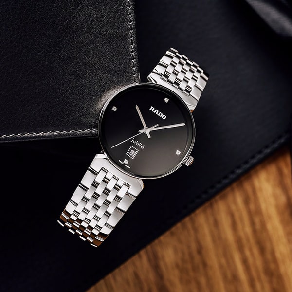 RADO © 2025 Eine stilvolle Armbanduhr aus der Rado Florence Kollektion mit silbernem Metallarmband und schwarzem rundem Zifferblatt mit dem Markennamen „Rado“. Die Uhr liegt auf einer schwarzen Lederoberfläche mit teilweise sichtbarem Holzhintergrund.