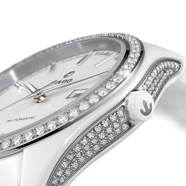 RADO © 2025 Nahaufnahme einer Luxusarmbanduhr aus der Rado Hyperchrome Kollektion mit einem eleganten weißen Design. Die Lünette und die Seiten der Uhr sind mit funkelnden Diamanten besetzt. Das Zifferblatt ist weiß mit silbernen Stundenmarkierungen und Zeigern. Die Uhr verfügt über ein automatisches Uhrwerk und eine elegante Krone.