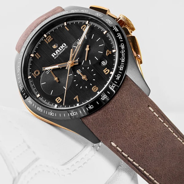 RADO © 2025 Eine Nahaufnahme einer Rado-Armbanduhr aus der Hyperchrome-Kollektion mit schwarzem Zifferblatt, goldenen Zeigern und Ziffern sowie einem Chronographen. Die Uhr verfügt über ein braunes Lederarmband mit sichtbaren Nähten und einer Metallschließe. Der weiße Hintergrund bietet einen klaren Kontrast zu der detailreichen Uhr.