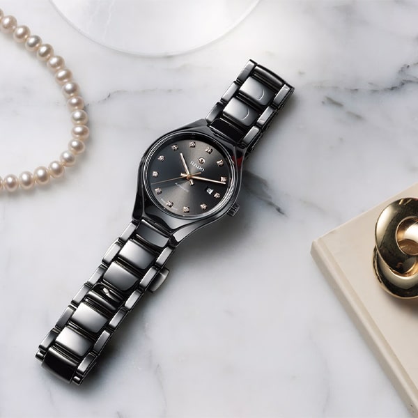 RADO © 2025 Eine elegante schwarze Armbanduhr aus der Rado True Round Kollektion mit dunklem Zifferblatt, das mit kleinen, funkelnden Stundenmarkierungen verziert ist. Die Uhr steht auf einer Marmoroberfläche neben einer Perlenkette und einem Teil eines Buches, in dessen Rahmen ein goldfarbener Stifthalter zu sehen ist.