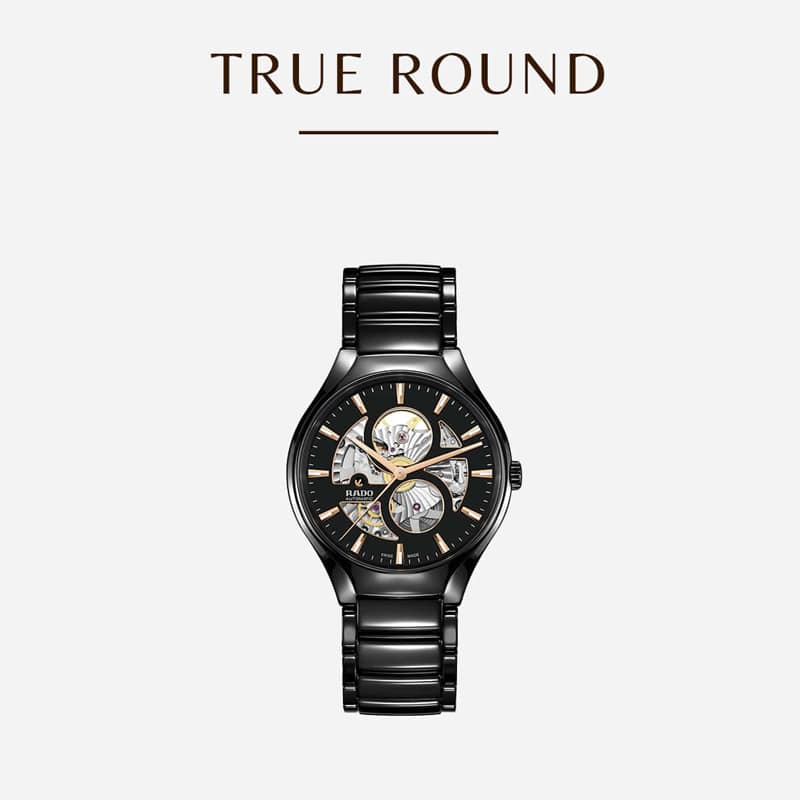 Eine elegante, schwarzmetallische Uhr mit sichtbaren Zahnrädern und goldenen Akzenten auf dem Zifferblatt. Oben im Bild steht der Text „TRUE ROUND“.