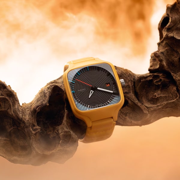 RADO © 2025 Eine moderne Armbanduhr aus der Rado True Square Kollektion verfügt über ein senfgelbes Armband und ein quadratisches Zifferblatt mit schwarzem Zifferblatt mit minimalistischen weißen Markierungen und rot-blauen Akzenten. Die Uhr ist stilvoll auf einem knorrigen Stück Treibholz mit einem warmen, nebelorangefarbenen Hintergrund platziert.