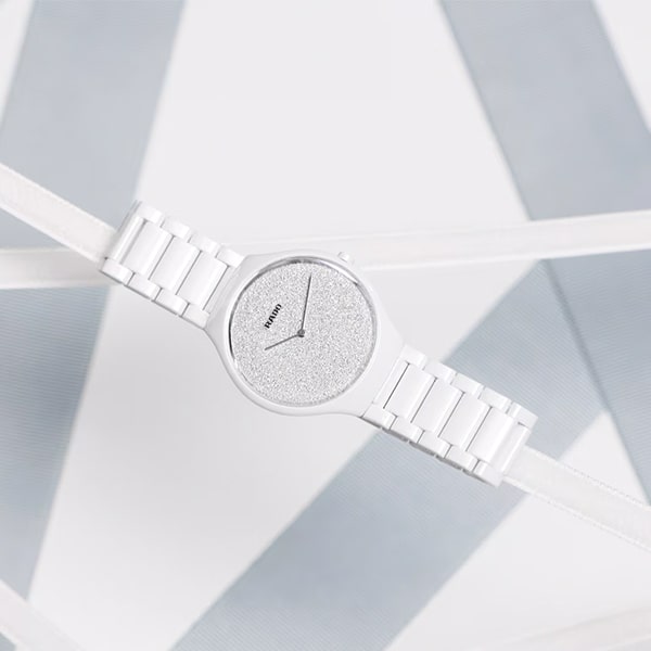 RADO © 2025 Eine stilvolle Armbanduhr aus der Rado True Thinline Kollektion mit weißem Armband und glitzerndem silbernem Zifferblatt wird vor einem hellen und schlichten Hintergrund mit geometrischen Mustern in Grau- und Weißtönen präsentiert. Die Uhr hat minimalistische Zeiger und keine sichtbaren Stundenmarkierungen, was ihr ein modernes, klares Design verleiht.