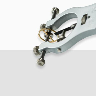 © Juwelier Wagner-Madler Nahaufnahme eines speziellen Uhrenlexikon-Werkzeugs, das ein kleines zahnradähnliches Bauteil auf einem weiß-grauen Hintergrund hält. Das Werkzeug scheint für Präzisionsarbeiten oder mechanische Einstellungen konzipiert zu sein.