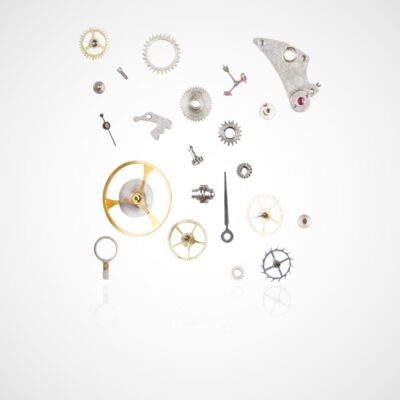© Juwelier Wagner-Madler Eine Sammlung verschiedener Uhrenkomponenten, darunter Zahnräder, Schrauben und eine Aufzugswelle, ist vor einem weißen Hintergrund angeordnet. Die Teile sind ausgebreitet und zeigen die komplizierten Elemente, aus denen der innere Mechanismus einer Uhr besteht. Es ähnelt einem zum Leben erweckten Uhrenlexikon.