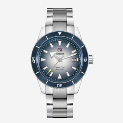 RADO © 2025 Eine silberne Armbanduhr mit Metallarmband und blauer Lünette. Das Zifferblatt ist grau mit weißen Stundenmarkierungen und Zeigern sowie einem Datumsfenster bei 3 Uhr. Unter der 12-Uhr-Markierung ist der Markenname „RADO“ zusammen mit „Captain Cook“ zu sehen, was die Uhr zu einem uhrenlexikonwürdigen Eintrag macht.
