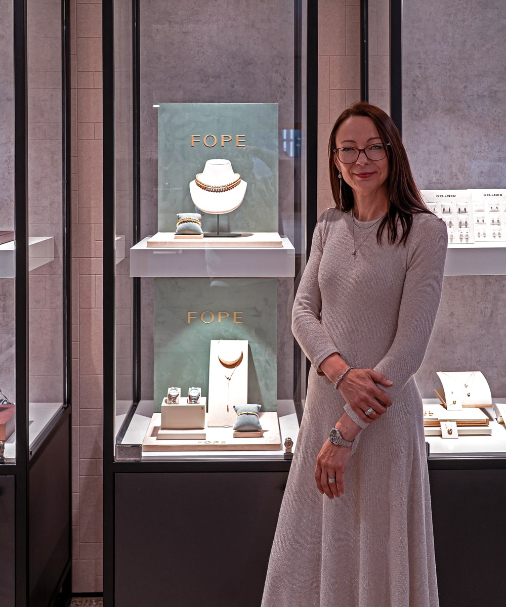 © Juwelier Wagner-Madler e.K. Eine Frau in einem langen, hellen Kleid steht vor einer Schmuckvitrine mit FOPE-Halsketten und -Armbändern im Atelier Wagner-Madler. Sie lächelt, trägt eine Brille und hat die Arme leicht verschränkt.