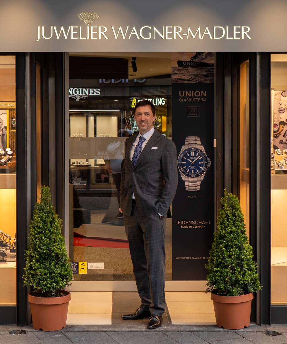 © Juwelier Wagner-Madler e.K. Ein Mann im Anzug steht am Eingang des Ateliers Wagner-Madler, einem Juweliergeschäft mit Topfpflanzen an der Tür und eleganten Uhrenauslagen im Inneren.
