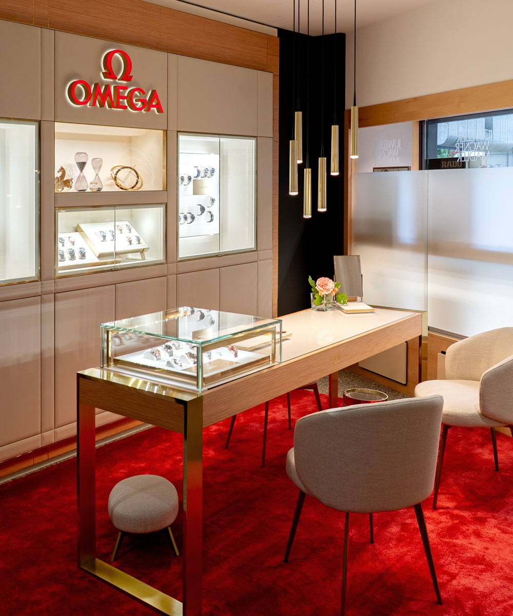 © Juwelier Wagner-Madler e.K. Die moderne Omega-Boutique von Atelier Wagner-Madler ist mit einem roten Teppich, Uhrenvitrinen, einem Holztisch mit Glasplatte, weißen Stühlen und Pendelleuchten ausgestattet, die eine elegante und einladende Atmosphäre schaffen.