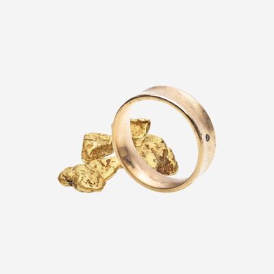 Ein einfacher Goldring liegt vor einem kleinen Haufen ungeschliffener Goldnuggets vor einem schlichten weißen Hintergrund. Der Ring ist leicht geneigt, wodurch seine glatte Oberfläche und runde Form zur Geltung kommen. Er erinnert an die Eleganz, die in jedem Schmucklexikon-Eintrag zu finden ist.