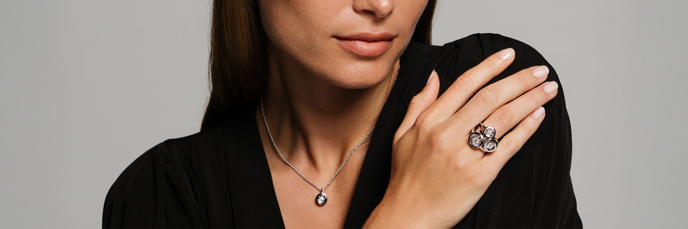 Eine Frau in einem schwarzen Oberteil trägt eine silberne Halskette mit einem runden Anhänger und einen großen Ring mit drei runden Steinen an ihrer Hand, die auf ihrer Schulter ruht - perfekt für jeden Schmucklexikon-Liebhaber.