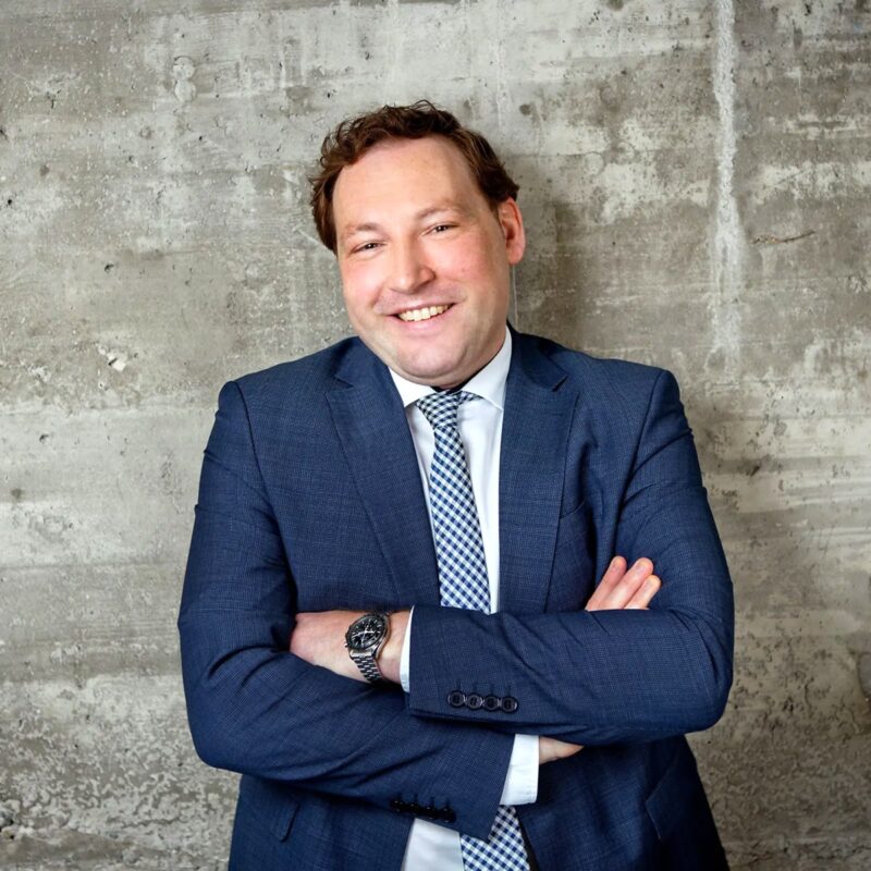 © Juwelier Wagner-Madler e.K. Ein Mann mit kurzen Haaren, einem blauen Anzug, weißem Hemd und karierter Krawatte steht lächelnd mit verschränkten Armen vor einer strukturierten Betonwand. Außerdem trägt er eine Uhr an seinem linken Handgelenk und strahlt Selbstvertrauen aus, als wäre er bereit, sein Team zu führen.