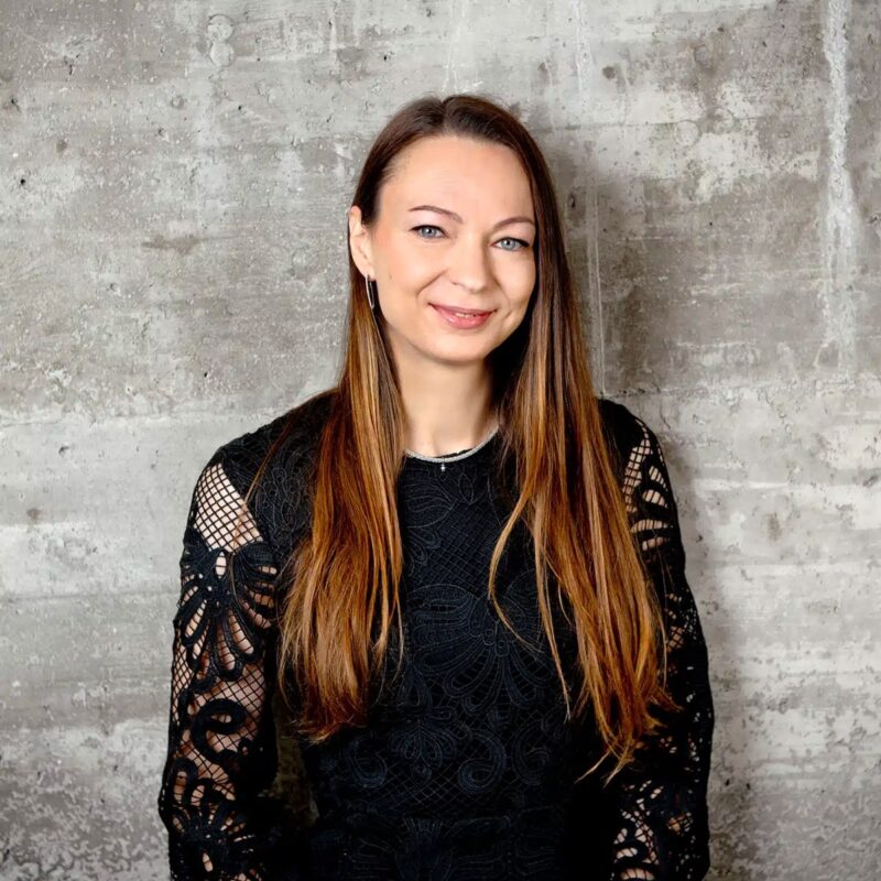 © Juwelier Wagner-Madler e.K. Eine Frau mit langen, glatten braunen Haaren lächelt in die Kamera. Sie trägt ein schwarzes, langärmliges Spitzentop. Der Hintergrund ist eine graue, strukturierte Wand, die ihren selbstbewussten Look ergänzt und sie ganz wie die Teamleiterin erscheinen lässt, die sie verkörpert.