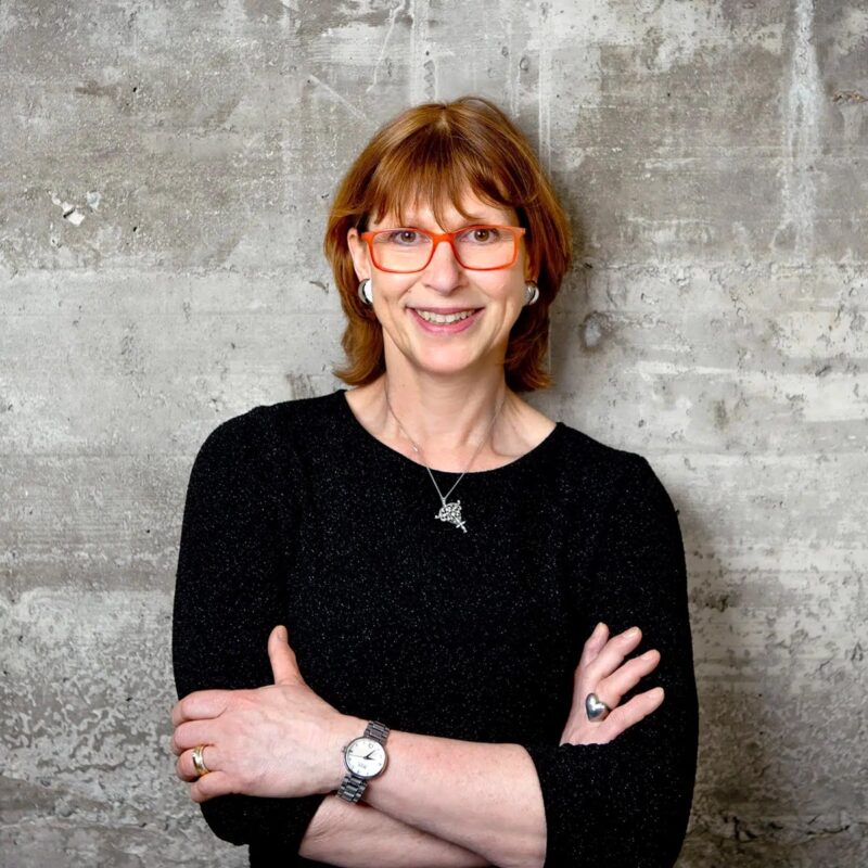 © Juwelier Wagner-Madler e.K. Eine Frau mit kurzen, rotbraunen Haaren und roter Brille steht mit verschränkten Armen vor einer Betonwand. Sie trägt ein schwarzes Oberteil, eine silberne Halskette und eine Uhr. Sie lächelt herzlich in die Kamera und strahlt Selbstvertrauen aus, als wäre sie bereit, jedes Team zum Erfolg zu führen.