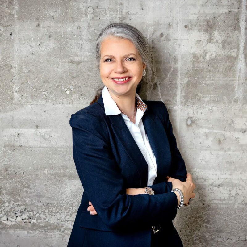 © Juwelier Wagner-Madler e.K. Eine Frau mit langen, grauen, zurückgebundenen Haaren steht lächelnd mit verschränkten Armen vor einer rauen Betonwand. Sie trägt einen marineblauen Blazer über einem weißen Hemd mit Knöpfen, was ihren professionellen Teamgeist widerspiegelt, und trägt nur wenige Accessoires.