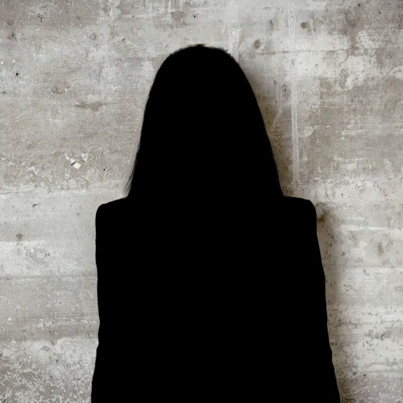 © Juwelier Wagner-Madler e.K. Die Silhouette einer Person mit langen Haaren und einem Blazer steht vor einer strukturierten, hellen Betonwand und erinnert an die stille Stärke eines Teammitglieds. Die Gesichtszüge der Person bleiben im Schatten verborgen.