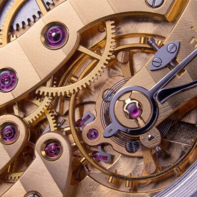 © Juwelier Wagner-Madler e.K. Das Uhrenlexikon zeigt anhand einer Nahaufnahme komplizierter Uhrzahnräder und -mechanismen verschiedene goldfarbene Zahnräder, Federn und Schrauben. In einigen Teilen sind winzige rosa Edelsteine eingelassen, die das komplexe Innenleben einer mechanischen Uhr veranschaulichen.
