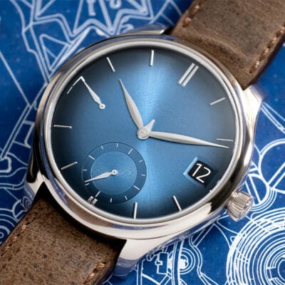 © Juwelier Wagner-Madler e.K. Eine Luxusarmbanduhr von Uhrenlexikon mit poliertem Edelstahlgehäuse, blauem Sunburst-Zifferblatt und braunem Lederarmband. Das Zifferblatt verfügt über silberne Zeiger, ein Datumsfenster auf der 3-Uhr-Position und Hilfszifferblätter. Die Uhr ist elegant auf einer blau gemusterten Oberfläche platziert.