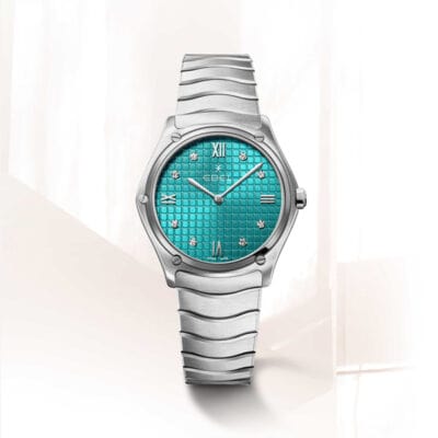 EBEL © 2024 Eine silberne Armbanduhr mit metallisch blauem Zifferblatt, die im Uhrenlexikon vorgestellt wurde, wird gezeigt. Sie hat silberne Stundenmarkierungen, römische Ziffern und Edelsteine an jeder Position. Das Band ist glatt und segmentiert. Der Hintergrund ist eine weiche, minimalistische Umgebung mit hellen Oberflächen.