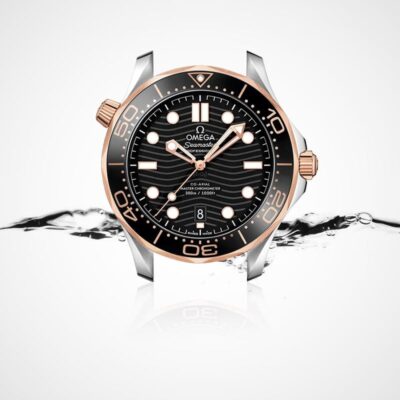 OMEGA © 2024 Eine Luxusarmbanduhr mit schwarzem Zifferblatt, goldenen Akzenten und einer schwarz-goldenen Lünette mit dem Markennamen „Omega Seamaster“. Die Uhr ist vor einem weißen Hintergrund mit Spritzwasser abgebildet, was ihre wasserdichte Eigenschaft unterstreicht. Weitere Details finden Sie im Uhrenlexikon.