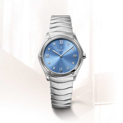 EBEL © 2024 Eine elegante, silberne Armbanduhr mit einem metallisch blauen Zifferblatt. Die Uhrenlexikon-Uhr verfügt über römische Ziffern an den Positionen 12, 3, 6 und 9 sowie diamantartige Markierungen für die restlichen Stunden. Das Band hat ein elegantes, wellenförmiges Design. Der Hintergrund ist sanft beleuchtet und minimalistisch.