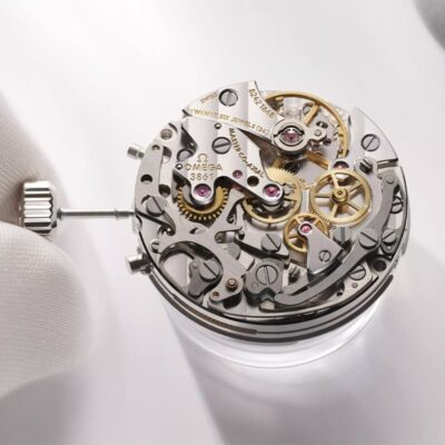 OMEGA © 2024 Nahaufnahme eines detaillierten Uhrwerks, das von einer Person mit weißen Handschuhen bedient wird. Die komplizierten Zahnräder, Zahnräder und Komponenten, einige mit der Aufschrift „OMEGA“ und „26 JEWELS“, sind sichtbar, während sie vom Experten anhand seines Uhrenlexikons sorgfältig freigelegt und eingestellt werden.