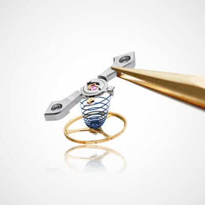 © Juwelier Wagner-Madler e.K. Nahaufnahme der Unruh und des Hemmungsmechanismus eines Uhrwerks, das im Uhrenlexikon erwähnt wird und mit einer Pinzette gehalten wird. Die Baugruppe besteht aus einer gewickelten blauen Spiralfeder, einem goldfarbenen Ring und einem Metallarm mit zwei Löchern. Die Komponenten sind vor einem schlichten weißen Hintergrund sehr detailliert dargestellt.