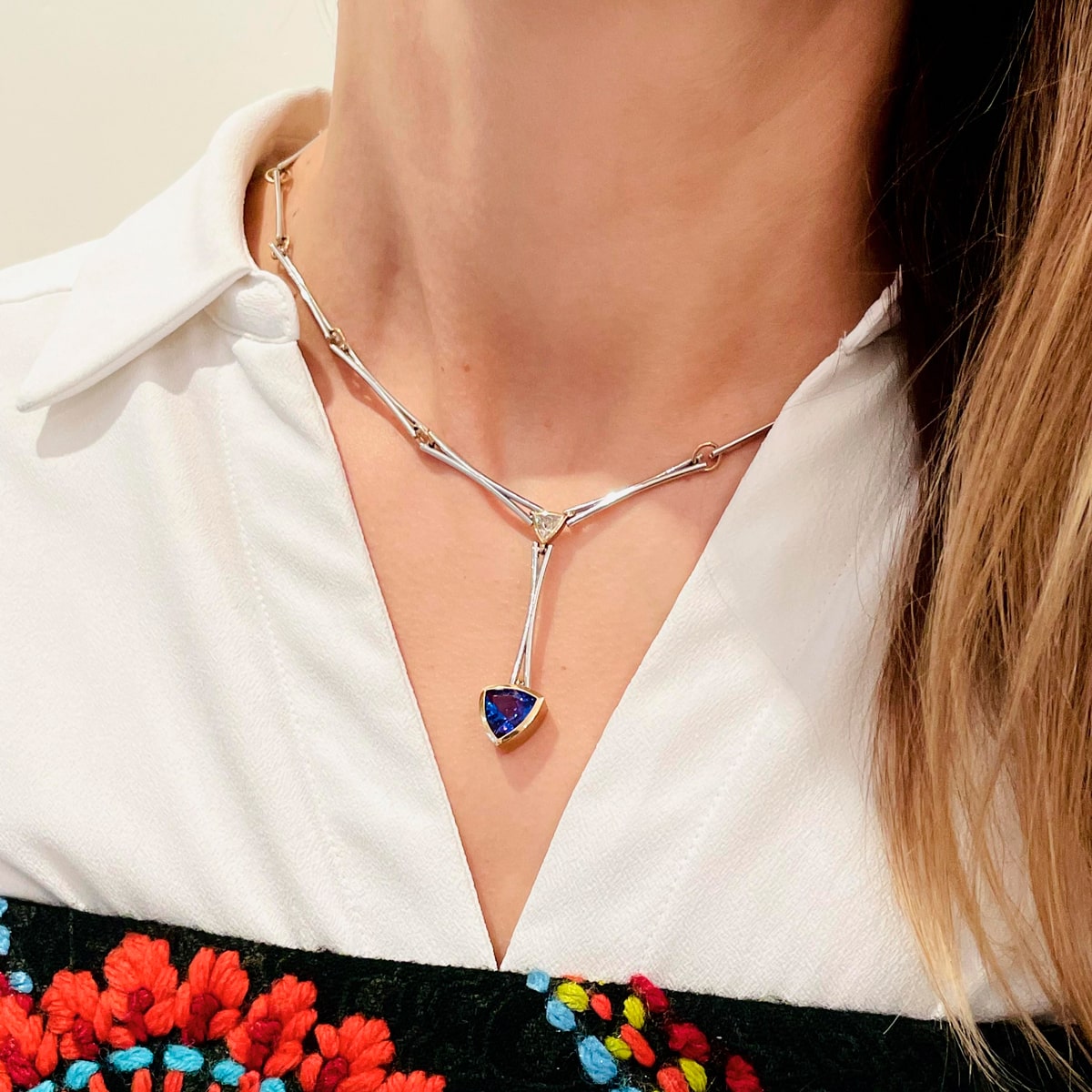 Eine Frau trägt ein weißes Hemd mit Kragen, ein schwarzes, rotes und blaues Oberteil mit Blumenstickerei von Atelier Wagner Madler und eine silberne Halskette mit einem geometrischen blauen Anhänger.