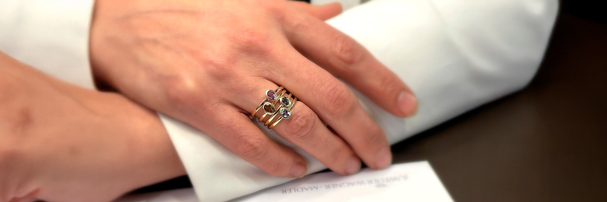 Nahaufnahme einer Hand, die einen Goldring mit drei Steinen des Ateliers Wagner Madler trägt, der sanft auf einer aufgerollten weißen Serviette neben einem teilweise sichtbaren Papier ruht.