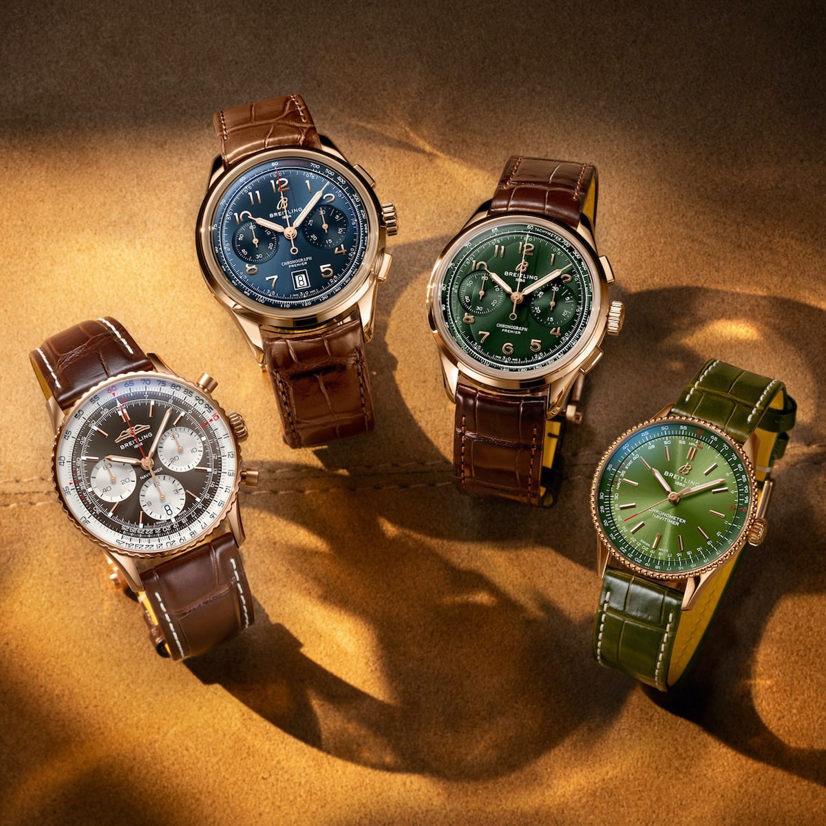 Vier luxuriöse Breitling-Armbanduhren mit Lederbändern sind ordentlich auf einer hellbraunen Fläche angeordnet. Die blauen, grünen und schwarzen Ziffernblätter mit goldenen oder silbernen Gehäusen harmonieren elegant mit braunen oder grünen Lederbändern. Schattierungen unterstreichen die raffinierte Darstellung.