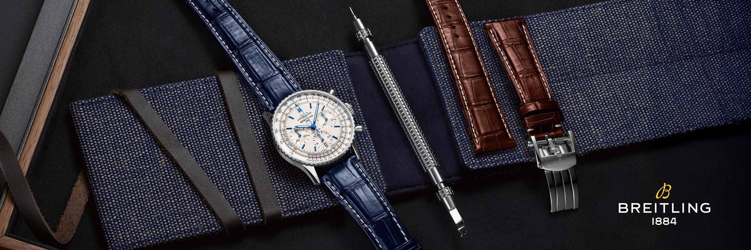 Eine luxuriöse Breitling-Uhr mit blauem Armband wird neben austauschbaren braunen Armbändern, einem Werkzeug und einer Metallschließe auf einem dunklen, strukturierten Hintergrund gezeigt. Das kultige Breitling-Logo und "1884" erscheinen in der Ecke.