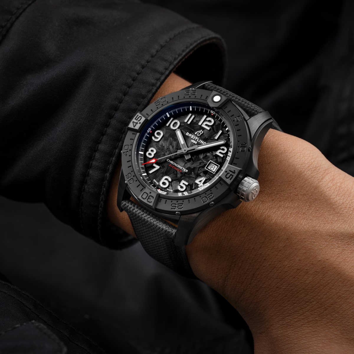 Eine Person trägt eine schwarze Breitling Avenger Armbanduhr mit einem großen Zifferblatt, weißen Ziffern und einem strukturierten schwarzen Armband. Der Zeiger und das Handgelenk sind vor einem schwarzen Ärmel abgebildet, der das kühne Design und die Details der Uhr hervorhebt.