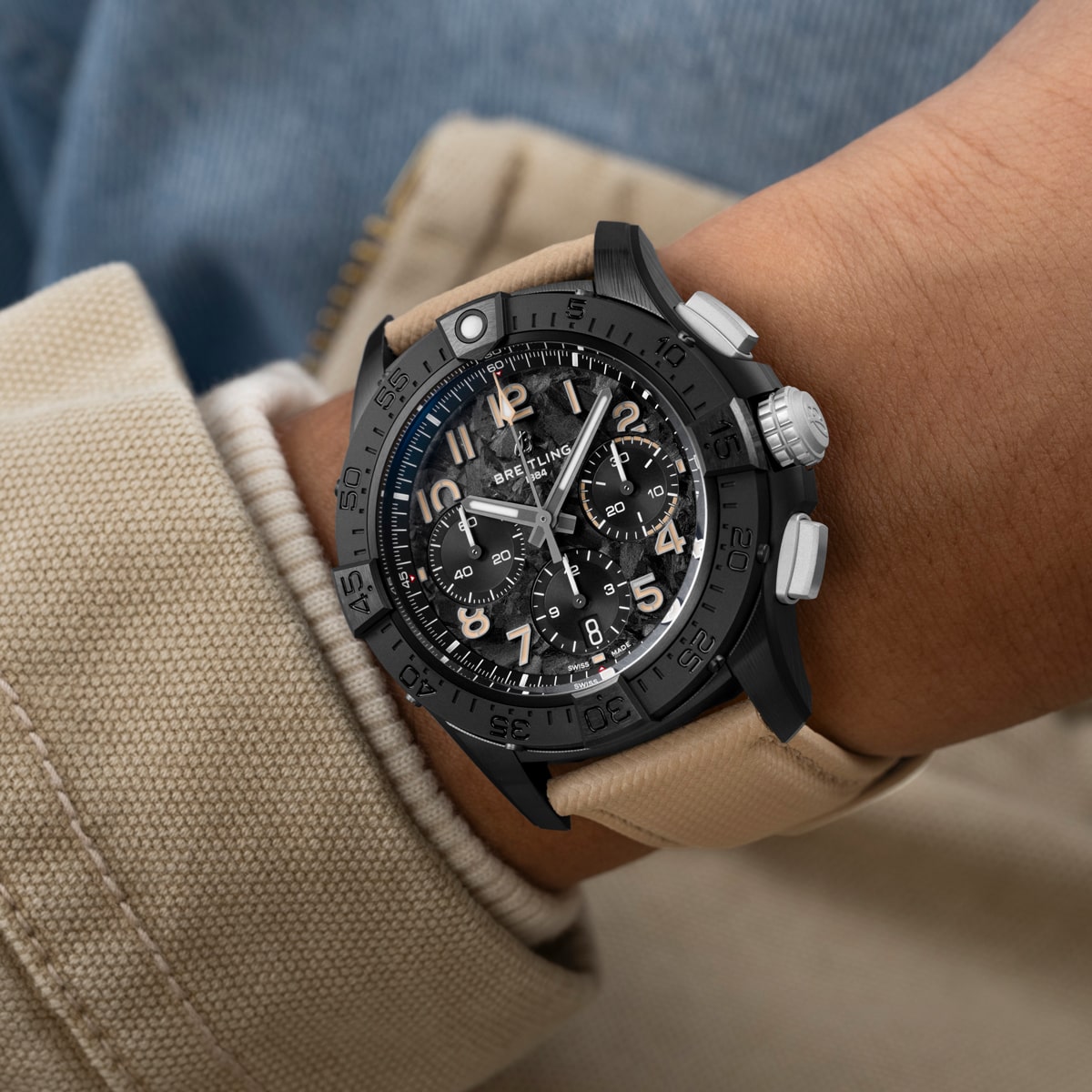 Nahaufnahme des Handgelenks einer Person, die eine große schwarze Breitling Avenger Chronographenuhr mit beigem Armband, schwarzem Zifferblatt und mehreren Unterzifferblättern über einer hellbraunen Jacke und blauen Jeans trägt.