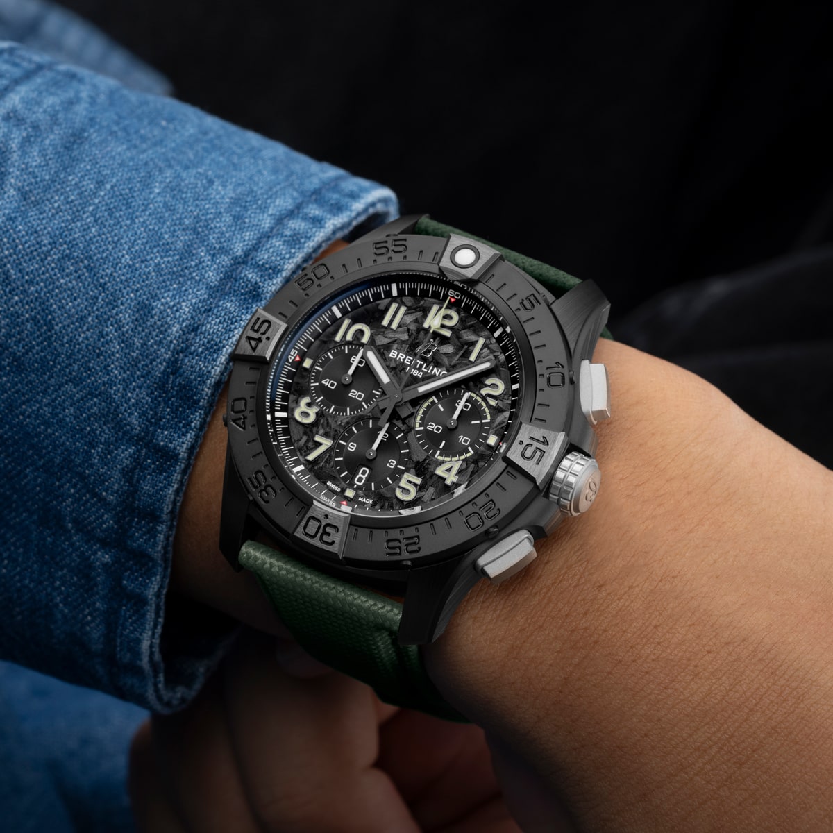 Eine Nahaufnahme des Handgelenks einer Person, die eine schwarze Breitling Avenger Chronographenuhr mit grünem Armband, silbernem Zifferblatt und weißen Ziffern trägt. Die Person ist mit einer blauen Jeansjacke bekleidet.