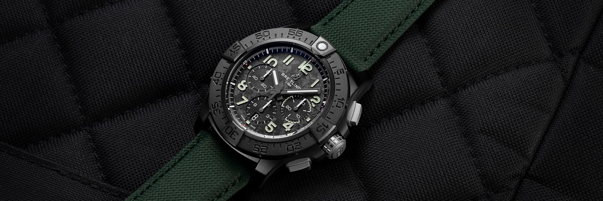 Eine schwarze Breitling Avenger Chronographen-Armbanduhr mit grünem Stoffarmband ruht auf einer strukturierten, schwarz gesteppten Oberfläche. Die Uhr hat fette Ziffern, mehrere Hilfszifferblätter und ein robustes Design.
