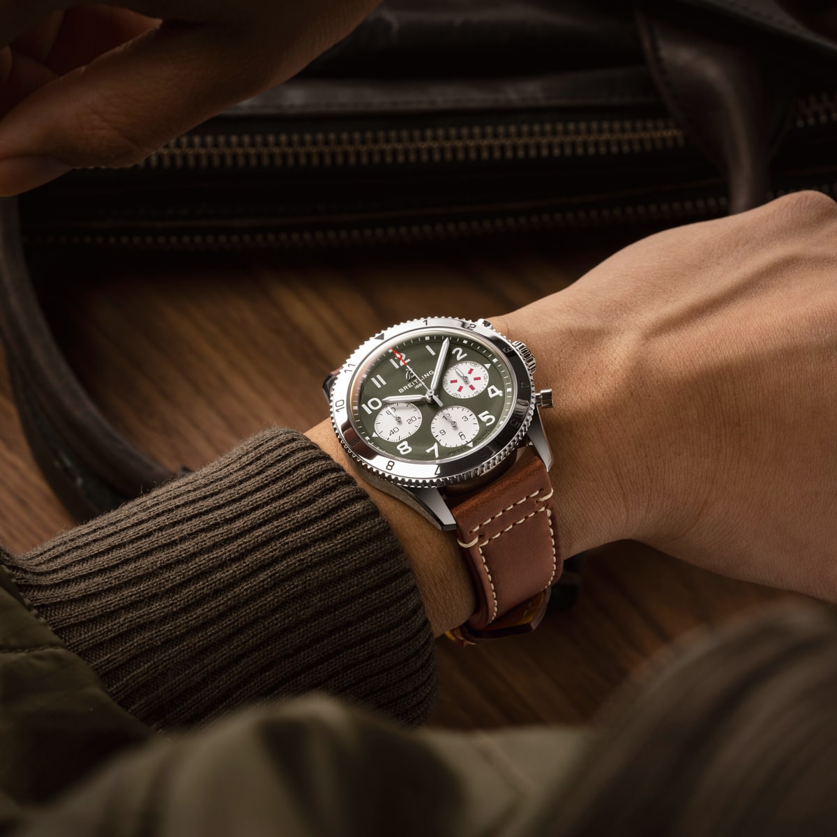 Eine Person trägt eine braune Breitling Classic AVI Armbanduhr mit Lederarmband und einem schwarz-weißen Zifferblatt mit mehreren Unterzifferblättern. Die Person ist mit einer braunen Jacke bekleidet und sitzt neben einer dunklen Ledertasche auf einer Holzfläche.