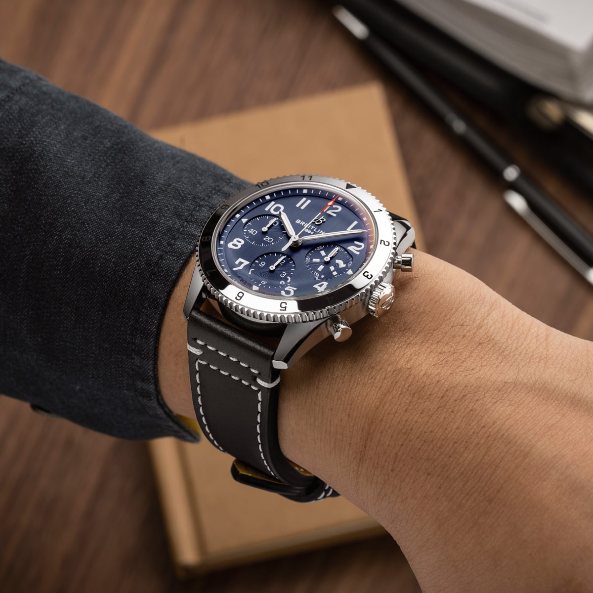 Eine Person, die eine Breitling Classic AVI Chronographen-Armbanduhr mit blauem Zifferblatt und schwarzem Lederarmband mit weißer Naht trägt, stützt ihren Arm auf einen Holztisch mit einem geschlossenen Notizbuch und einem Stift in der Nähe.