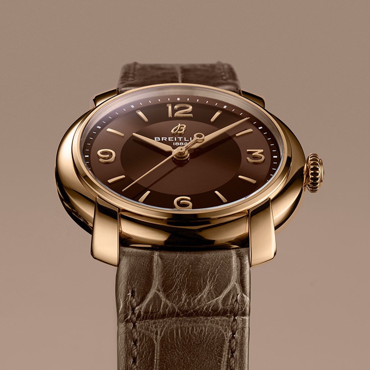 Eine Nahaufnahme einer Breitling-Armbanduhr mit braunem Zifferblatt, goldenem Gehäuse und braunem Lederarmband vor einem passenden beigen Hintergrund. Die Uhr zeigt die Zeit 10:09 an.