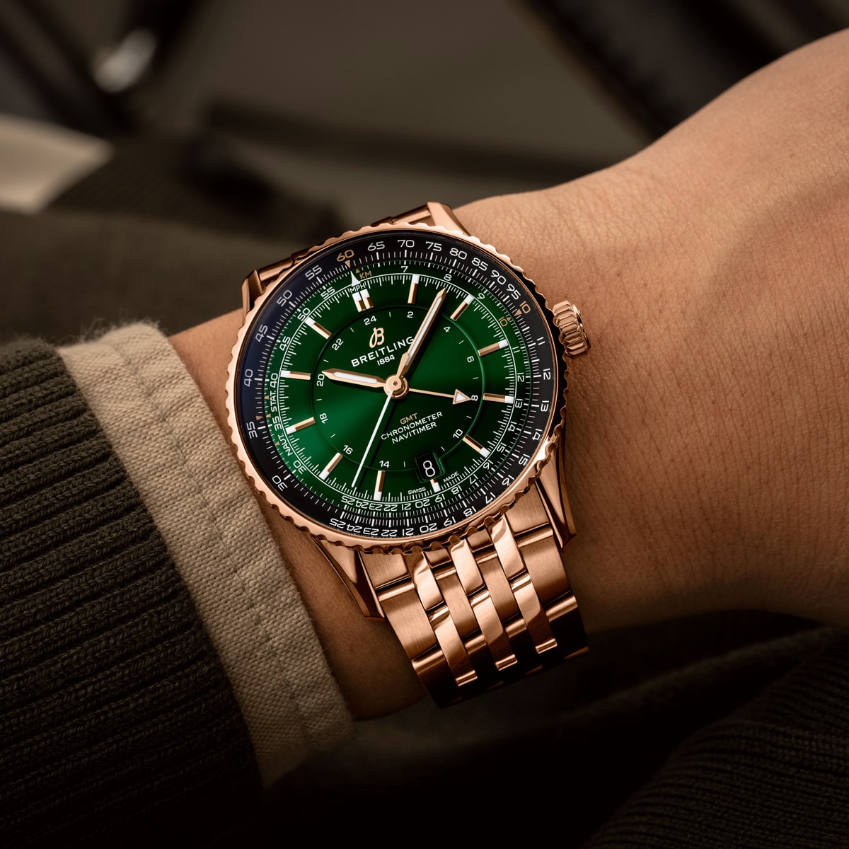 Eine Person trägt eine goldene Breitling Navitimer mit grünem Zifferblatt und Metallgliederarmband. Die Uhr verfügt über mehrere Zifferblätter und Markierungen, die perfekt zu ihrem dunklen, strukturierten Pullover passen.