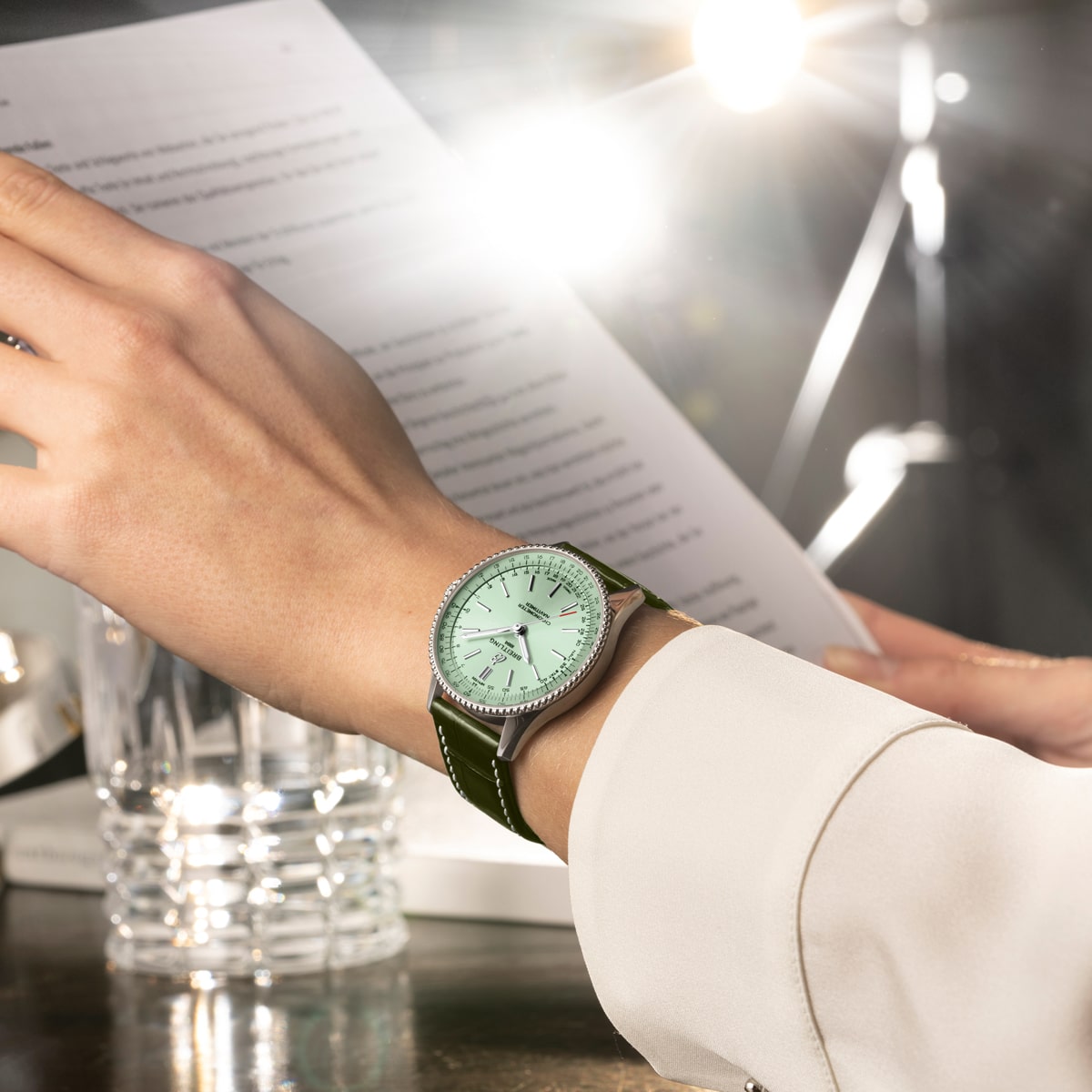 Eine Person, die eine grüne Breitling Navitimer-Armbanduhr trägt, hält ein Dokument. Im Hintergrund sind ein Glas Wasser und helles Studiolicht zu sehen. Die Person ist mit einem hellen, langärmeligen Hemd bekleidet.