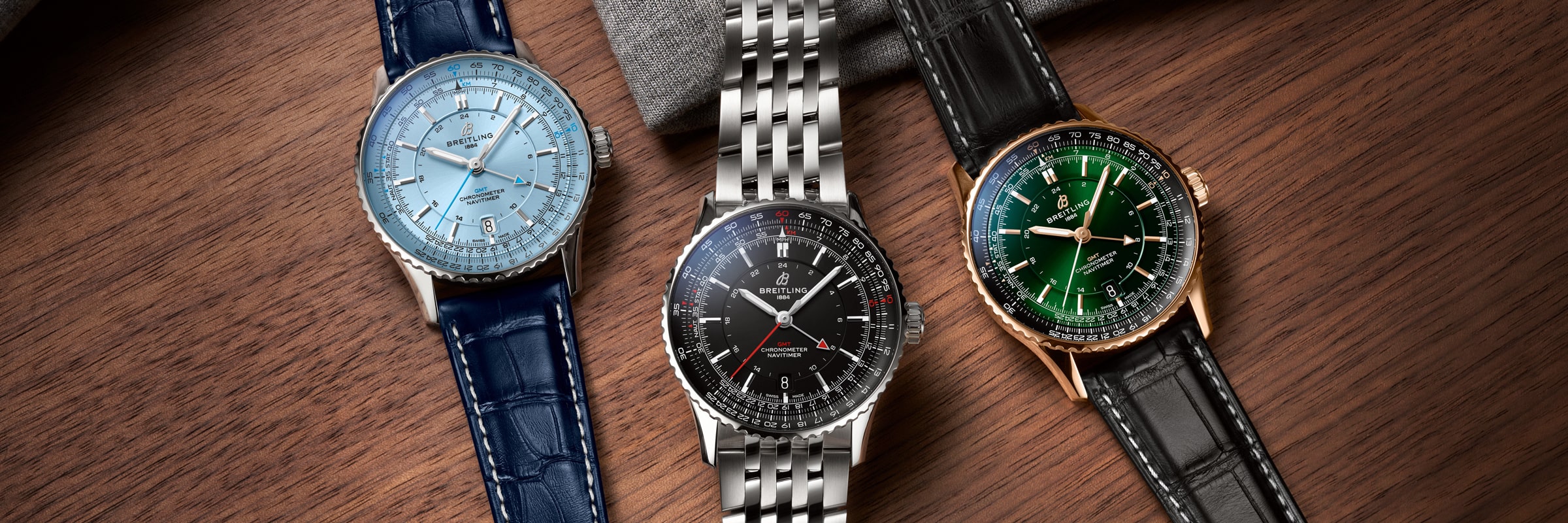 Drei luxuriöse Breitling-Armbanduhren, darunter eine markante Breitling Navitimer, sind auf einer Holzfläche ausgestellt: eine mit blauem Lederarmband und hellblauem Zifferblatt, eine mit Metallarmband und schwarzem Zifferblatt und eine mit schwarzem Lederarmband und grünem Zifferblatt.