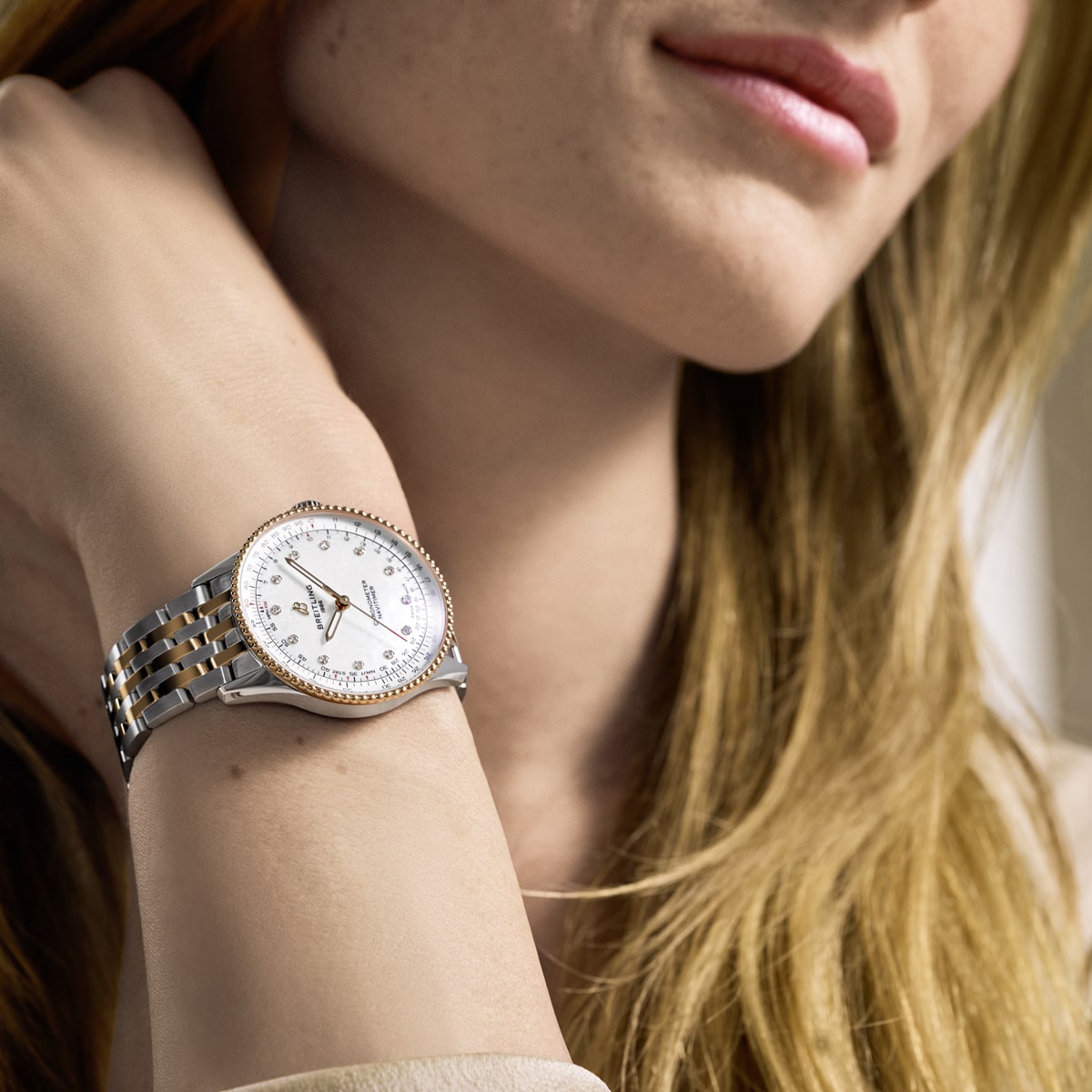 Eine Frau mit langem blondem Haar, die ihr Kinn auf die Hand stützt, präsentiert eine wunderschöne Breitling-Armbanduhr in Silber und Gold mit weißem Zifferblatt und Diamantindexen an ihrem Handgelenk.