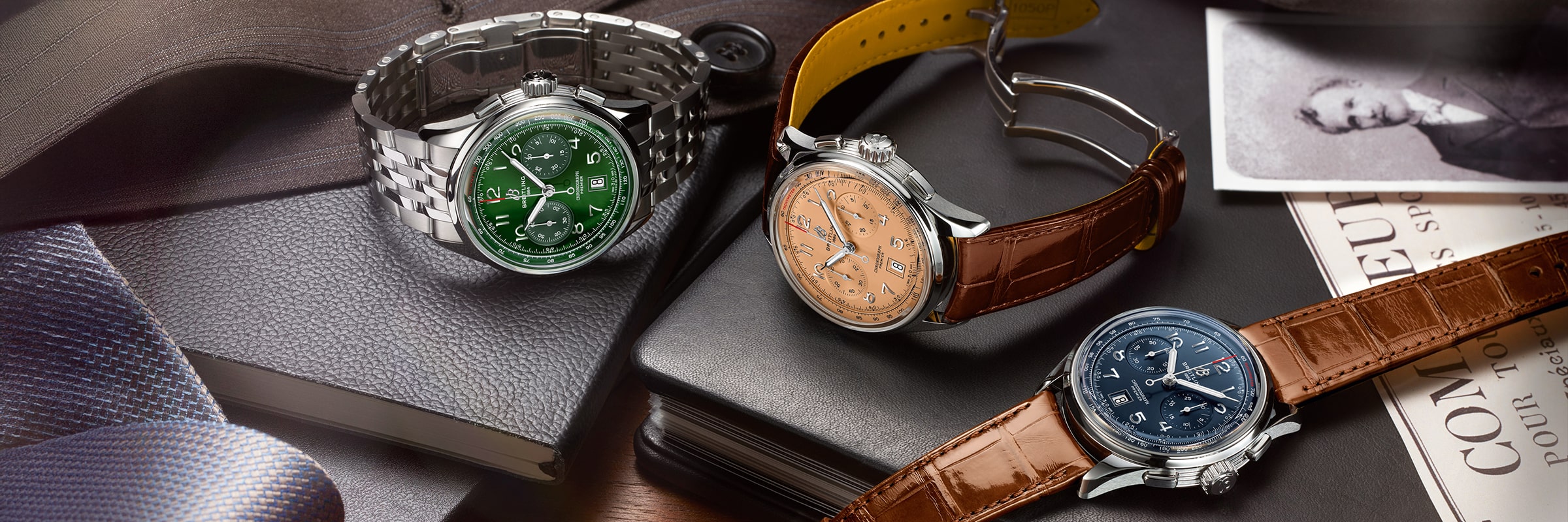 Drei luxuriöse Armbanduhren der Breitling Premier Kollektion mit grünen, lachsfarbenen und blauen Zifferblättern werden auf einem Tisch mit Lederarmbändern, einem Metallarmband, Notizbüchern, einer Krawatte und alten Schwarz-Weiß-Fotos präsentiert.