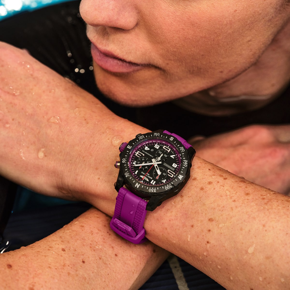 Eine Person mit heller Haut und feuchten Sommersprossen stützt ihre Arme ab und zeigt eine schwarze Sportuhr mit lila Armband aus der Breitling Professional Kollektion. Auf dem Zifferblatt sind mehrere Ziffernblätter und Markierungen zu sehen.