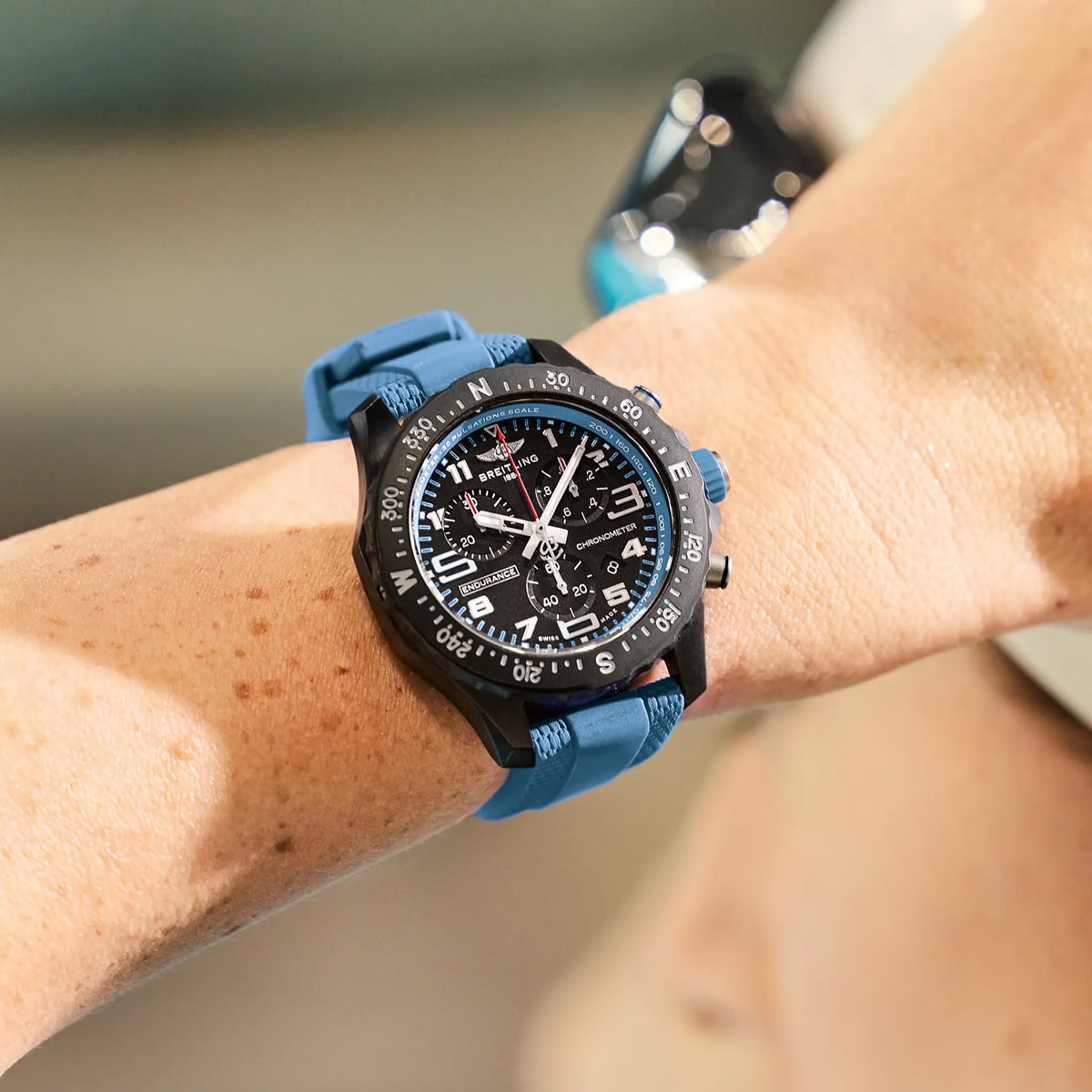 Nahaufnahme eines Handgelenks mit einer massigen schwarzen analog-digitalen Sportuhr mit blauem Armband, deren detaillierte Zifferblätter die Präzision der Breitling Professional Kollektion widerspiegeln. Der Hintergrund ist unscharf.