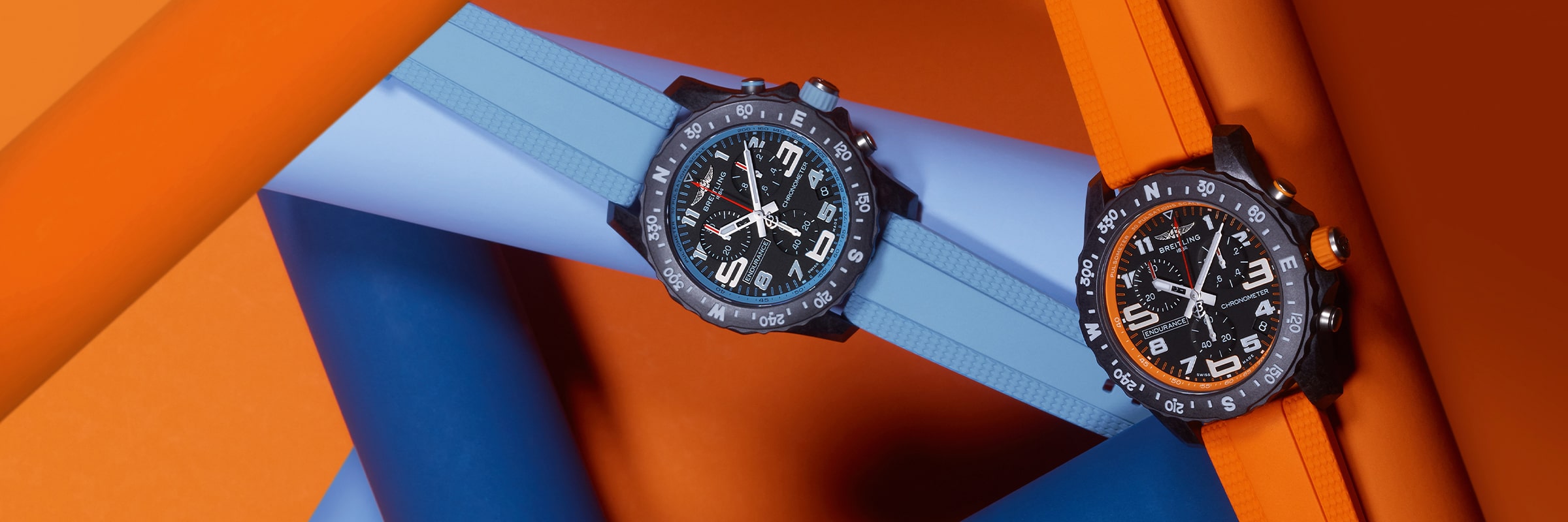 Zwei schwarze Uhren aus der Breitling Professional Kollektion mit großen Zifferblättern und fetten Ziffern ruhen auf ineinander verschlungenen orangefarbenen und blauen Röhren. Die eine hat ein blaues, die andere ein orangefarbenes Armband, das vor einem farblich passenden Hintergrund steht.